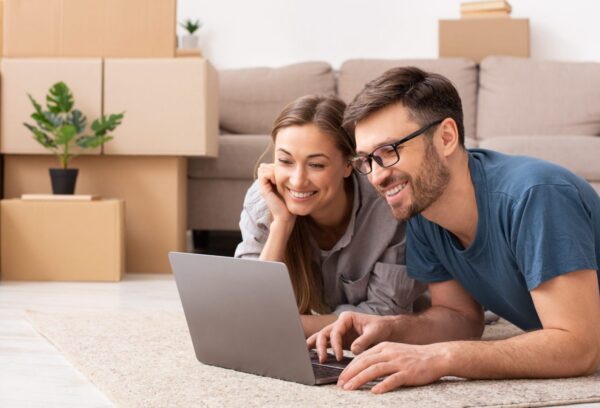 Happy couple searching online with a laptop over a cardboard box sitting on the floor while moving apartment, panorama, copy space
